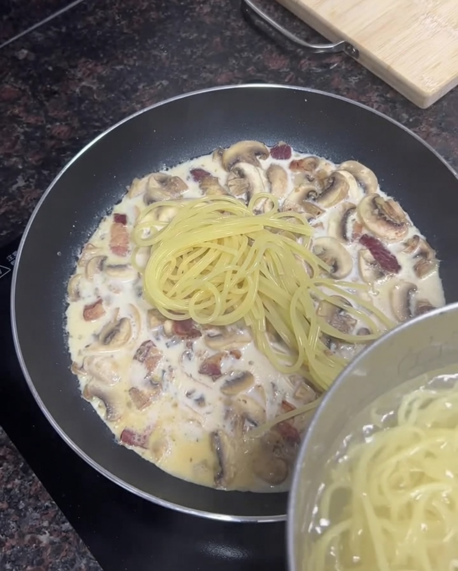 Spaghetti cotti aggiunti da una pentola in una padella con salsa cremosa al bacon e funghi.
