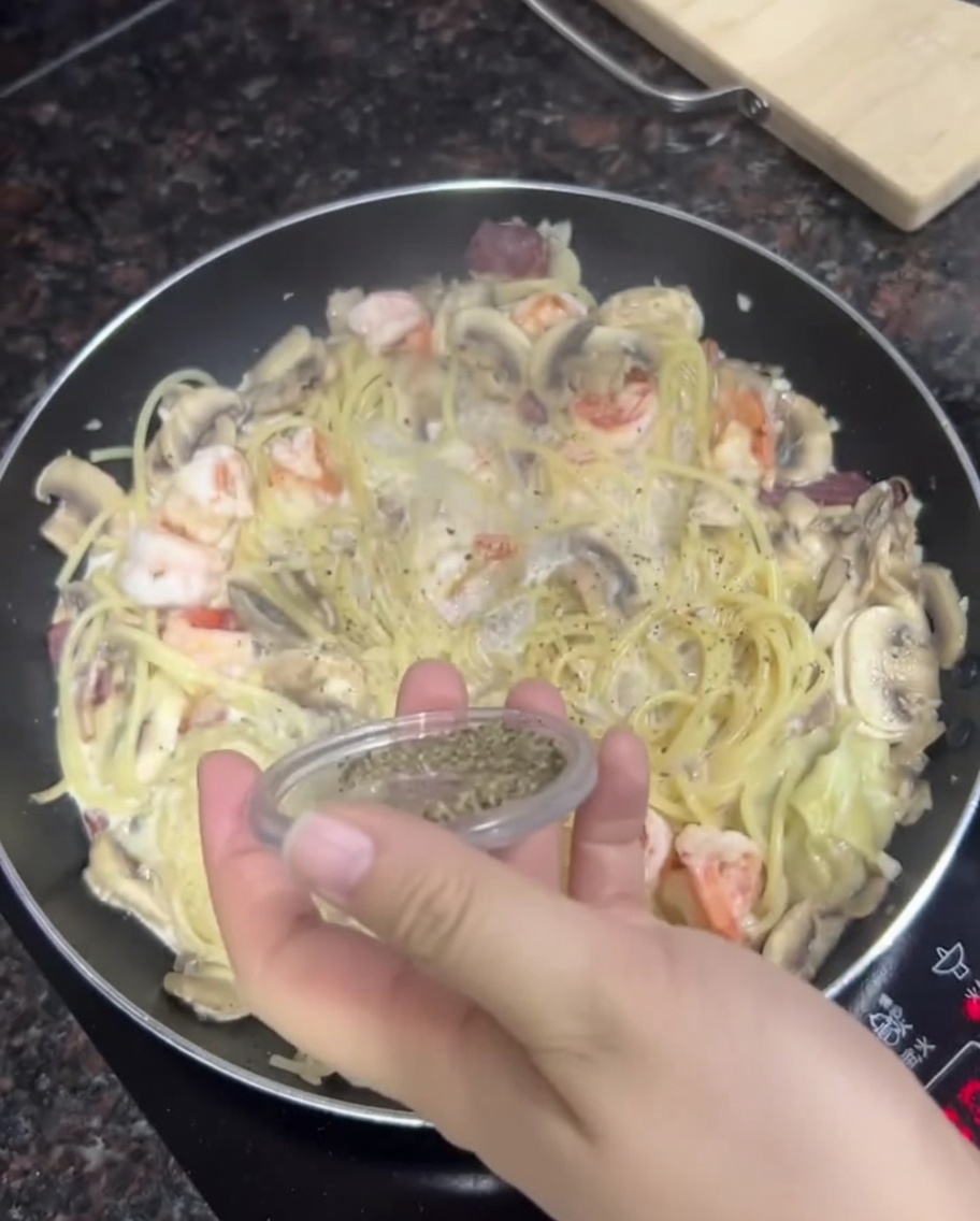 Una mano che spolvera pepe nero fresco sopra una padella di pasta cremosa.