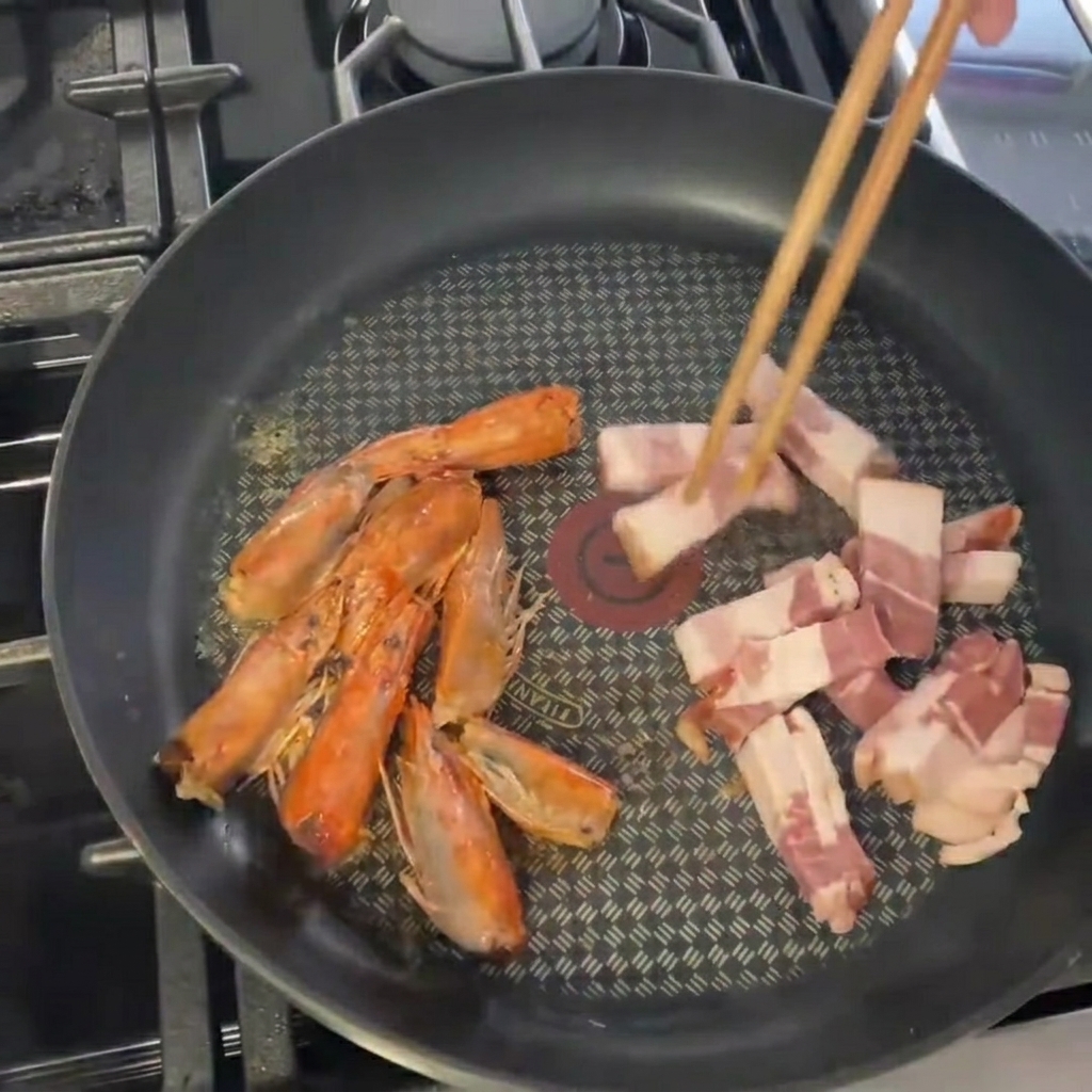 Pezzi di pancetta affettata aggiunti in una padella contenente già le teste di gamberi saltate.