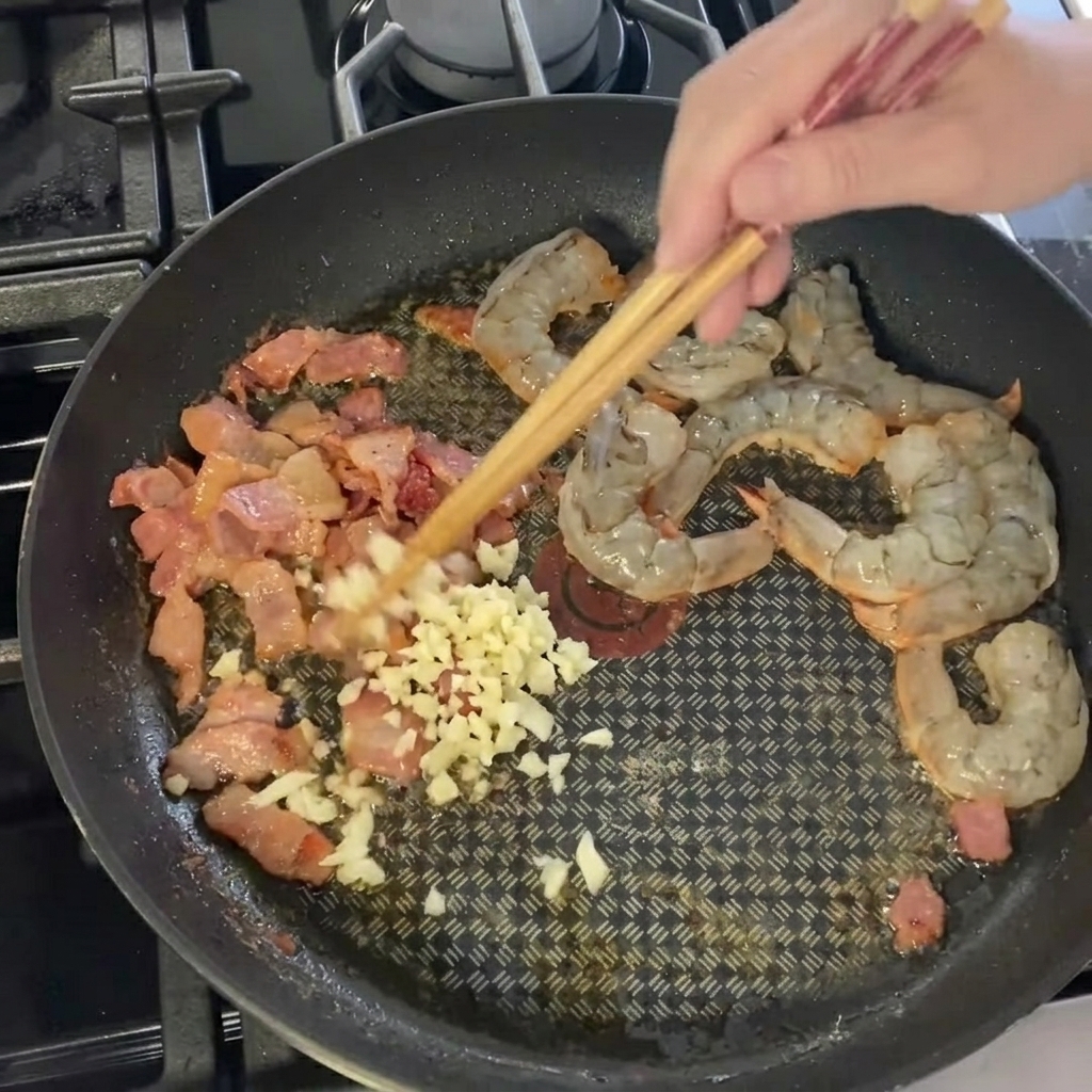 Aglio finemente tritato sparso sul composto di gamberi e pancetta in padella.