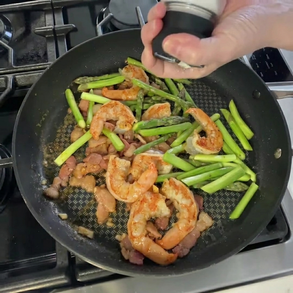 Mano che condisce il composto di gamberi e asparagi in padella con sale e pepe.
