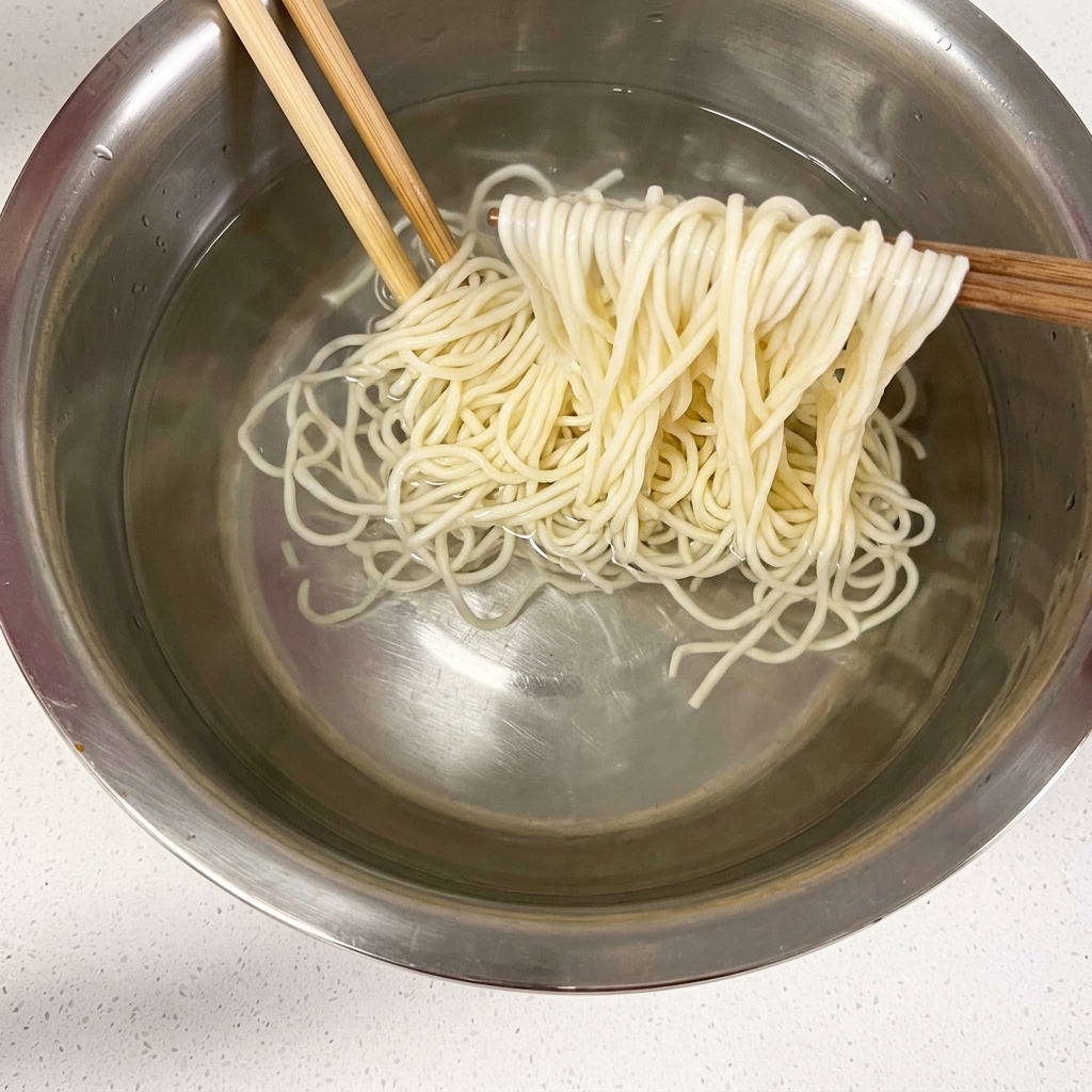 Noodles cotti sollevati con le bacchette sopra una ciotola di acqua fredda.
