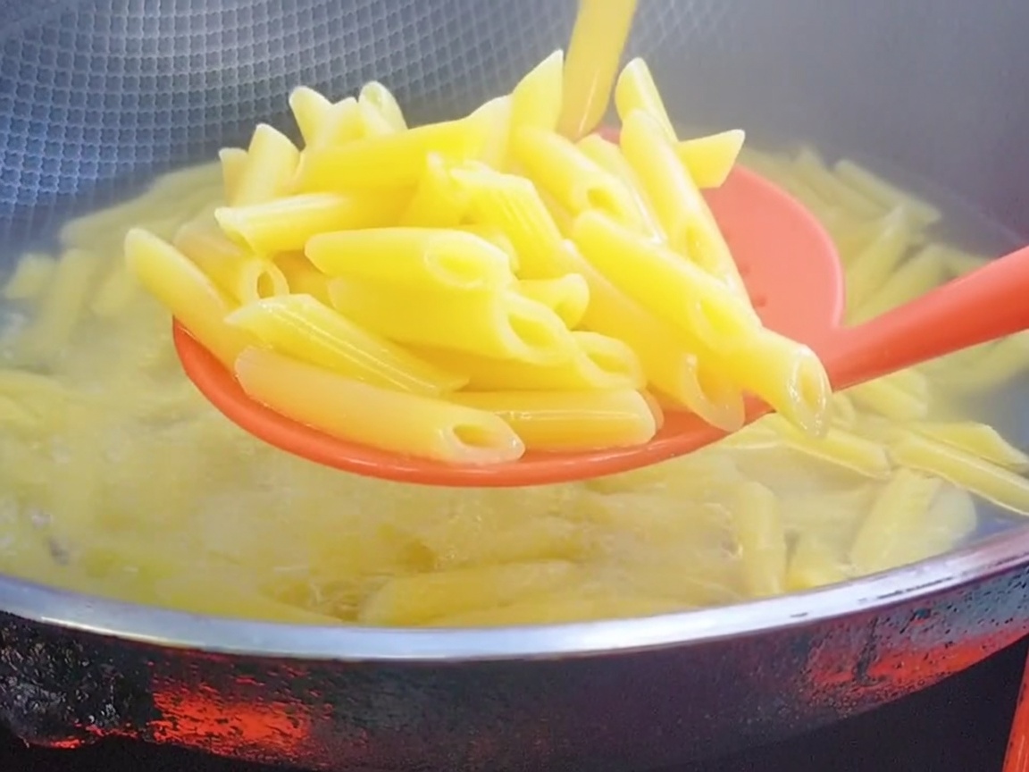 Un cucchiaio forato in silicone rosso che solleva le penne cotte dallacqua bollente.