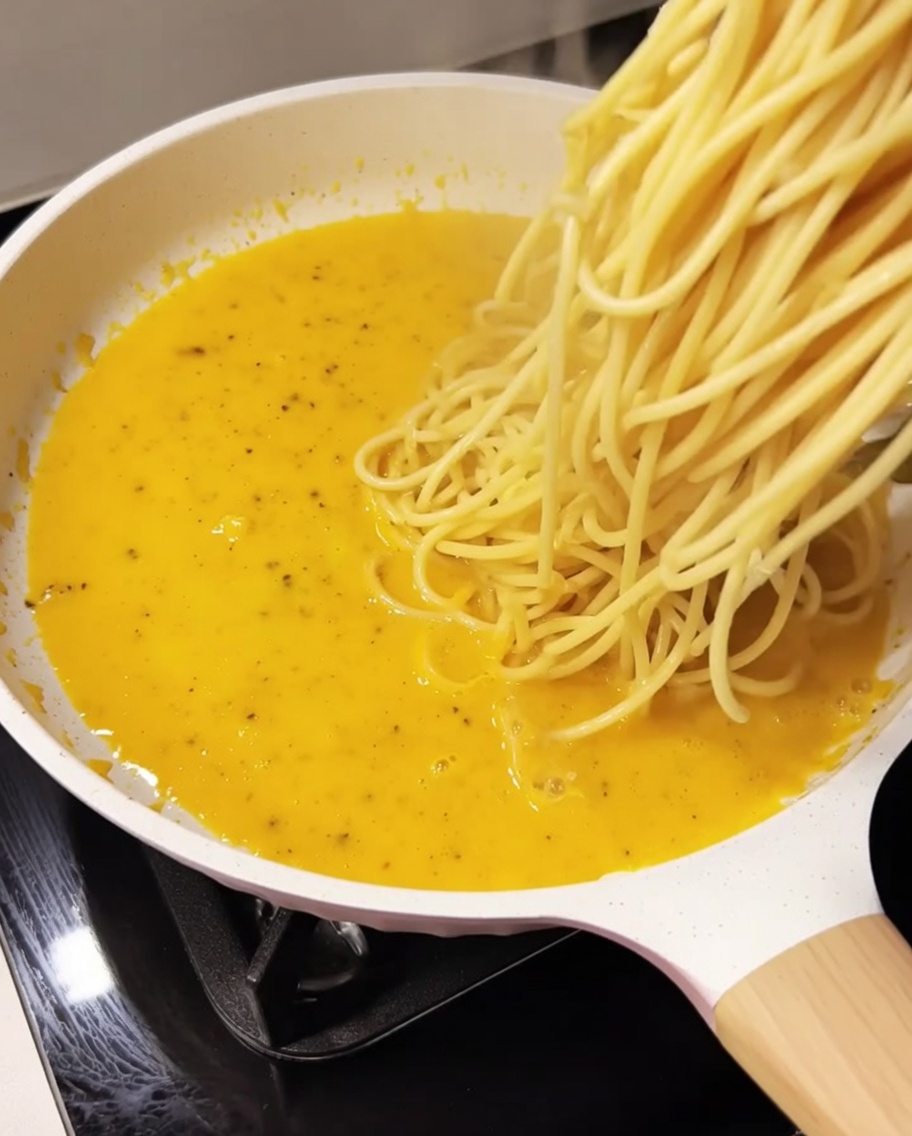 Spaghetti cotti uniti alla salsa gialla e cremosa in una padella bianca.