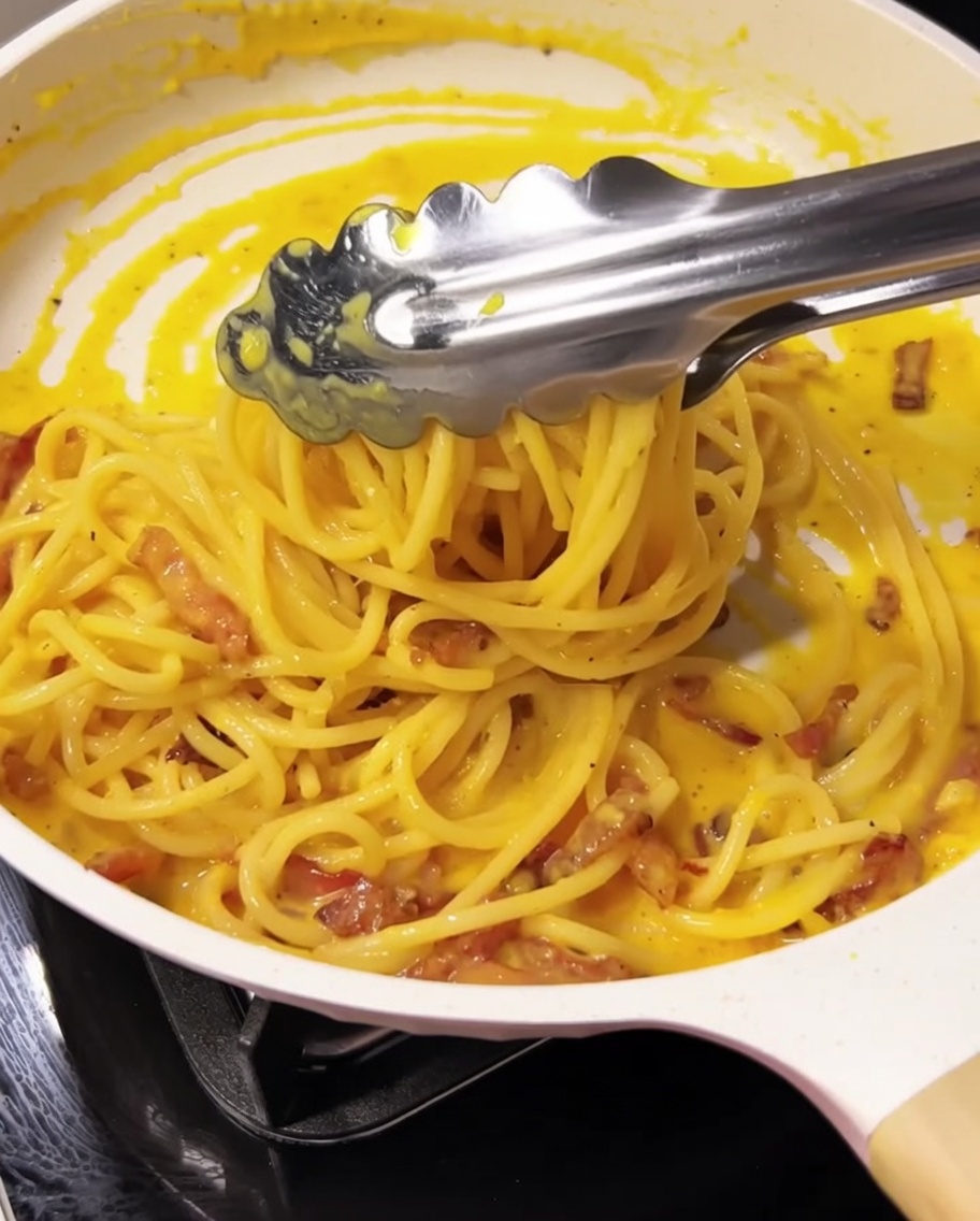 Pinze che mescolano gli spaghetti con la crema e il guanciale in padella.