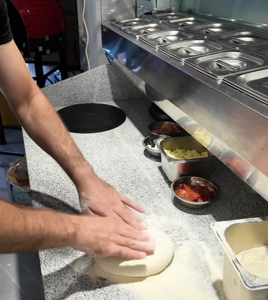 Mani che stendono una pallina di impasto per pizza su un piano di lavoro infarinato.