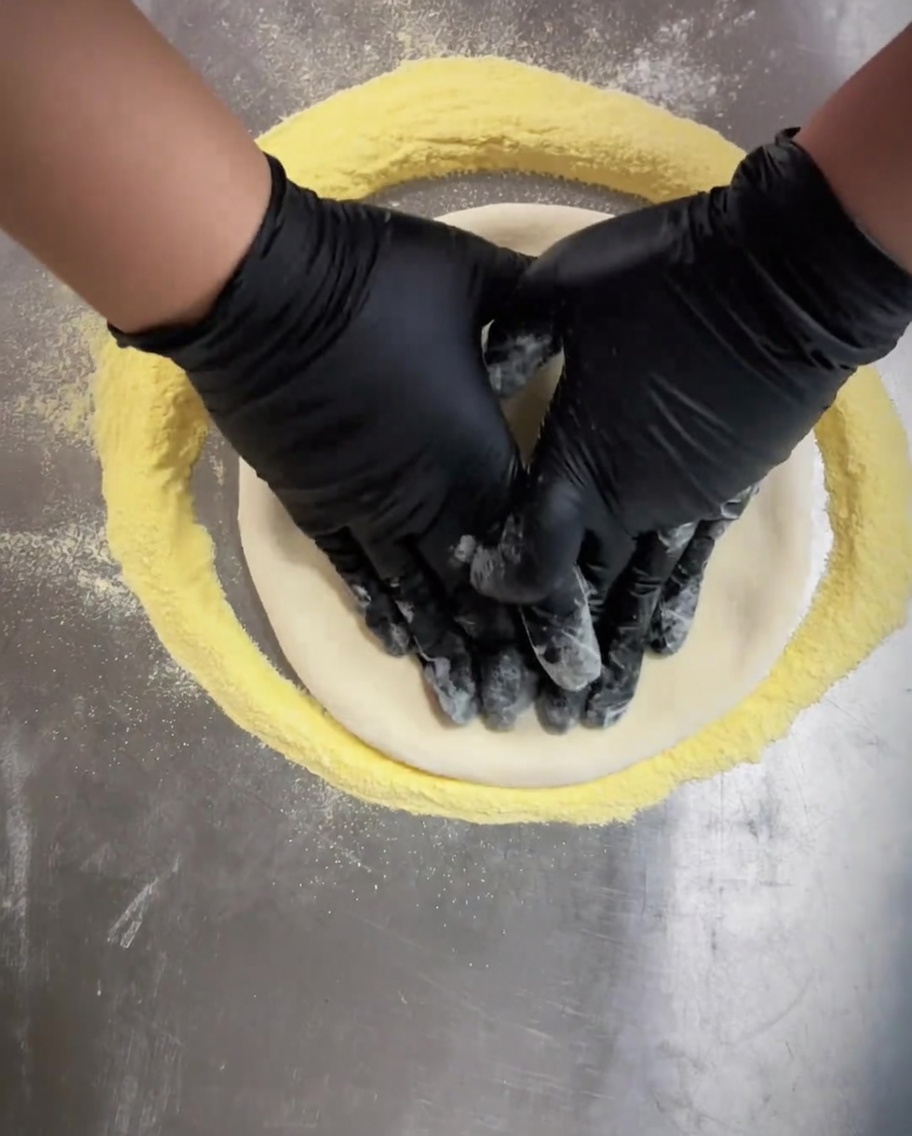 Mani con guanti neri che premono al centro dellimpasto della pizza per appiattirlo uniformemente.