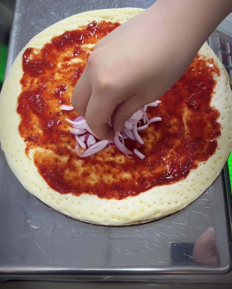 Pezzi di cipolla rossa e un denso strato di mozzarella grattugiata aggiunti sulla pizza.