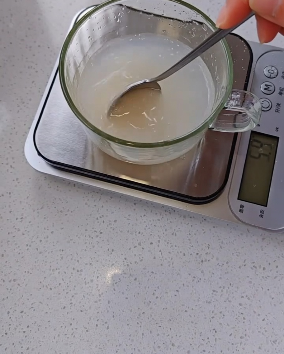 Una mano usa un cucchiaino per mescolare una miscela torbida di lievito e acqua in un misurino graduato trasparente su una bilancia da cucina.