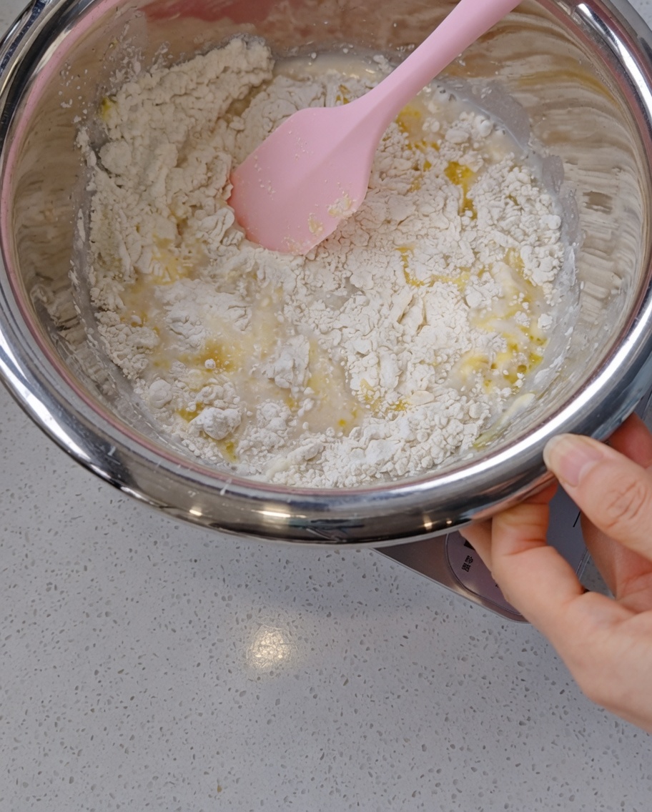 Una mano usa una spatola in silicone rosa per mescolare un impasto grezzo e umido con tracce dolio in una ciotola dacciaio.