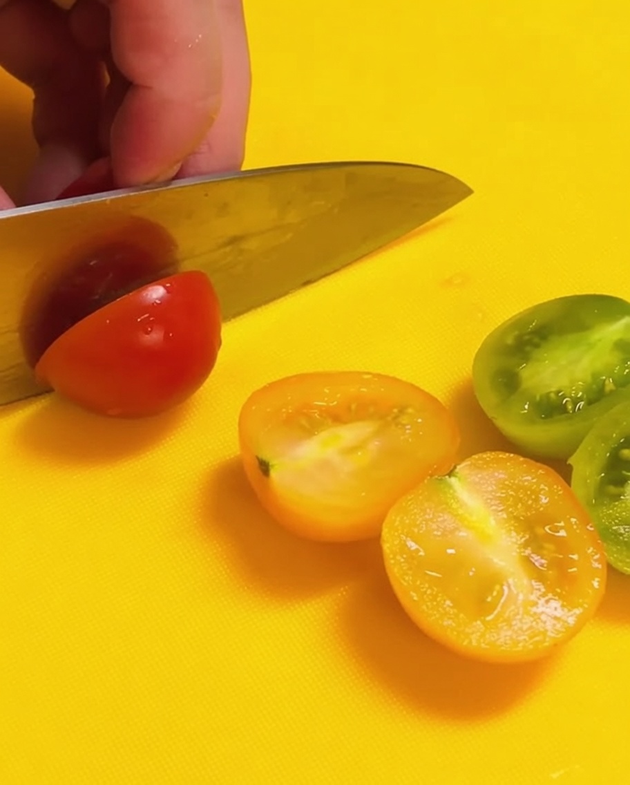 Un coltello taglia a metà dei pomodorini rossi e verdi su un tagliere giallo brillante.