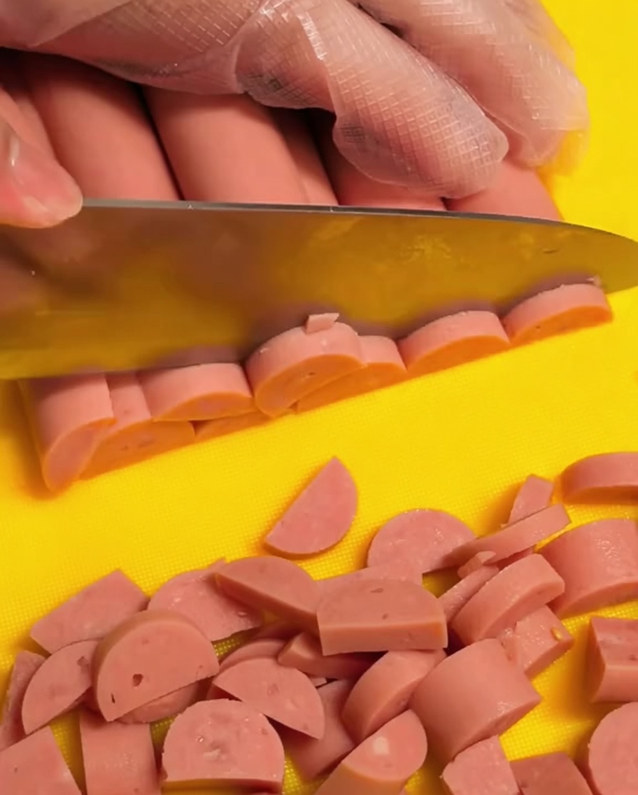 Una mano con guanto trasparente affetta dei wurstel di prosciutto a mezzaluna su un tagliere giallo.
