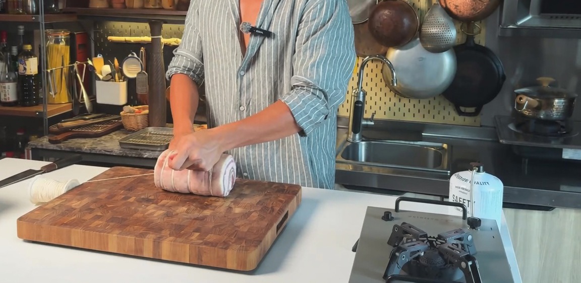 Una pancetta di maiale cruda arrotolata legata orizzontalmente con spago da cucina su un tagliere di legno.