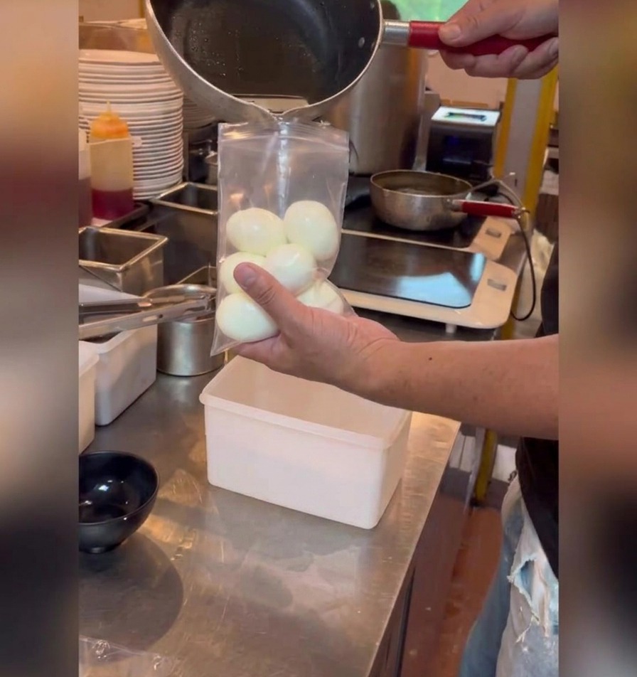 Una marinata scura a base di soia viene versata da un pentolino in un sacchetto di plastica trasparente pieno di uova barzotte sgusciate.