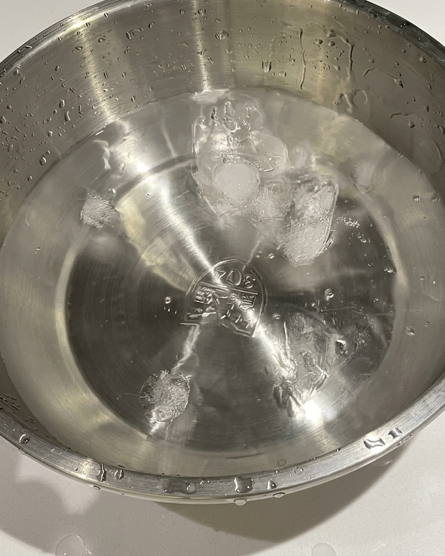 Uova cotte trasferite da una pentola in una ciotola di metallo con acqua fredda e cubetti di ghiaccio.