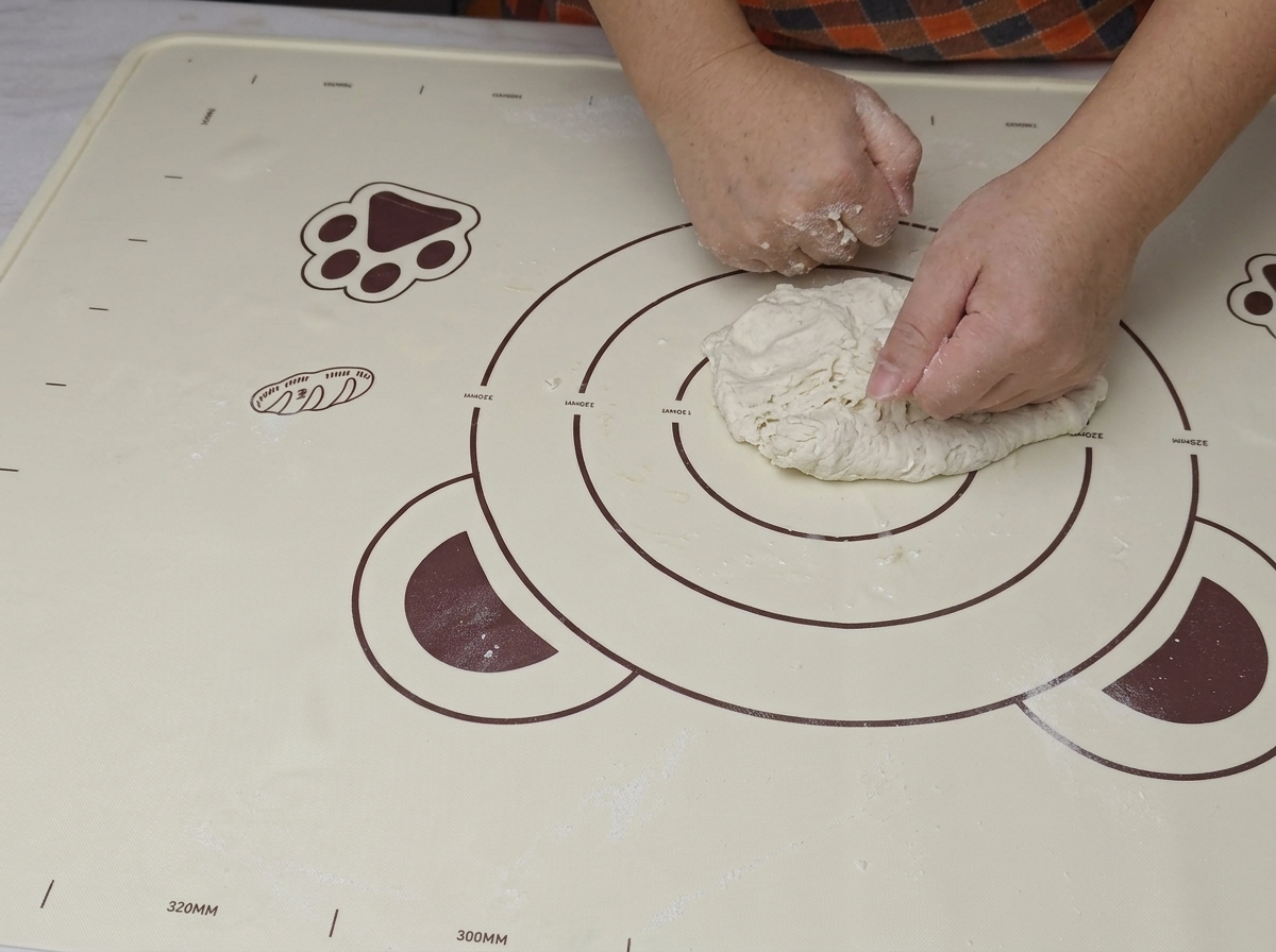 Due mani che premono e ripiegano una palla di impasto grezzo su un tappetino in silicone.