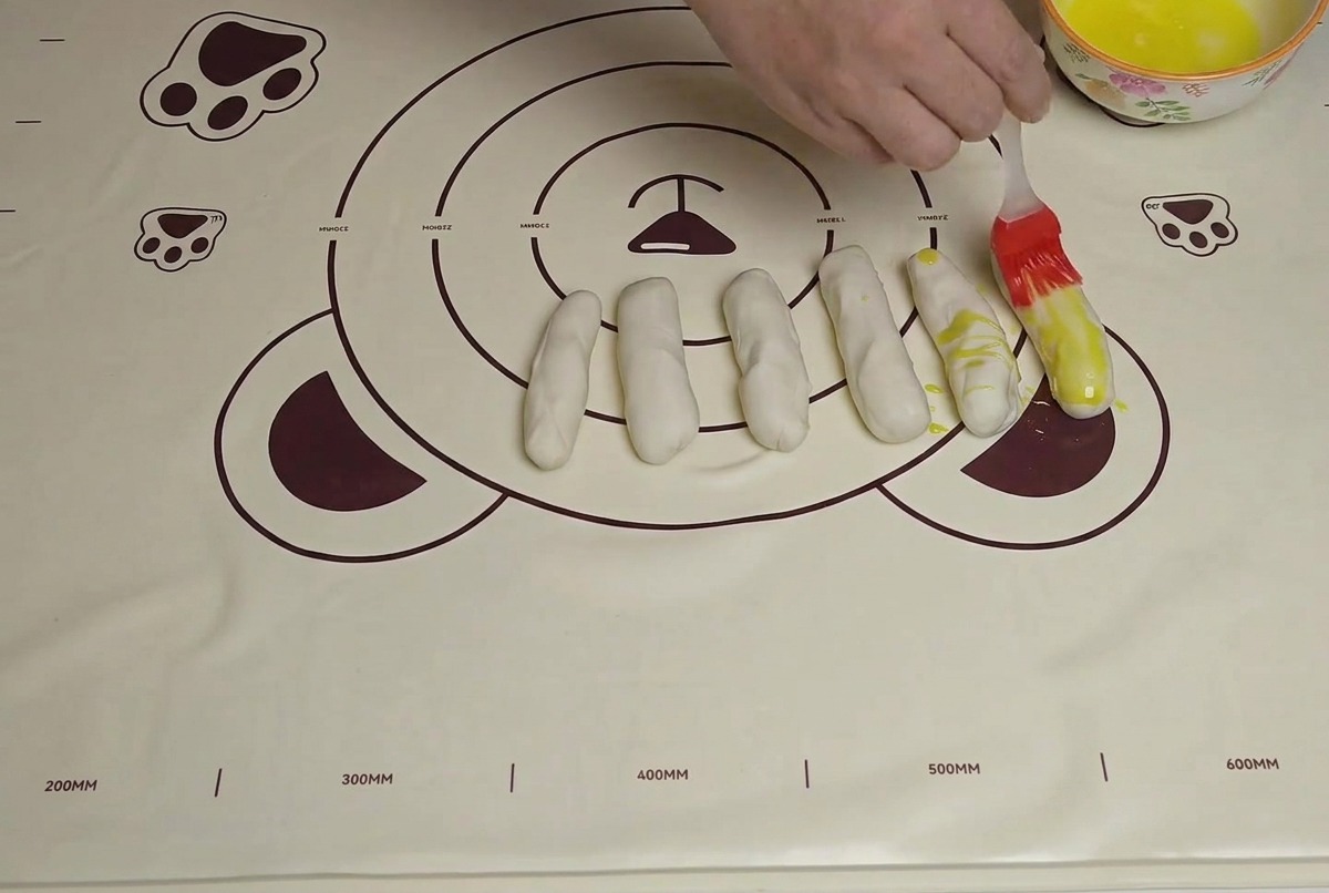Strisce di impasto su un tappetino mentre vengono spennellate con olio con un pennello in silicone.