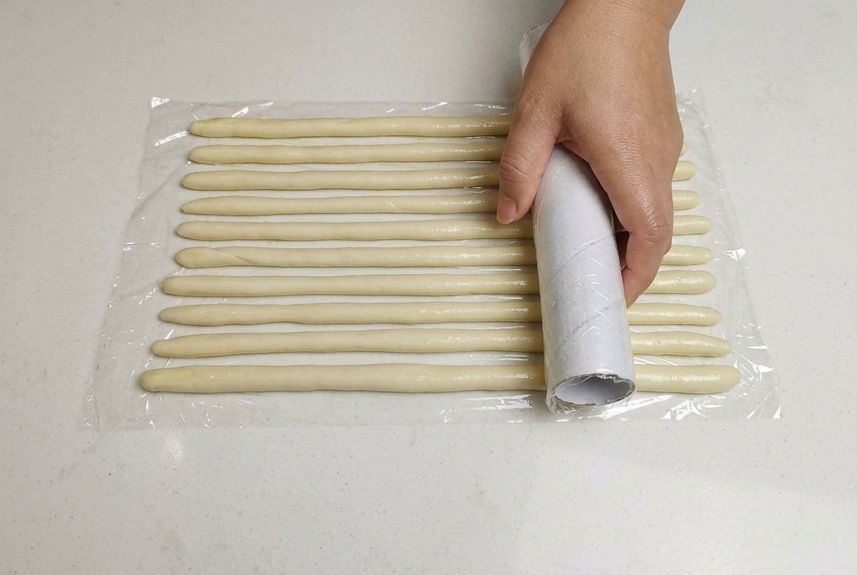 Una mano che copre con pellicola trasparente dei cordoni di impasto disposti su una superficie bianca.