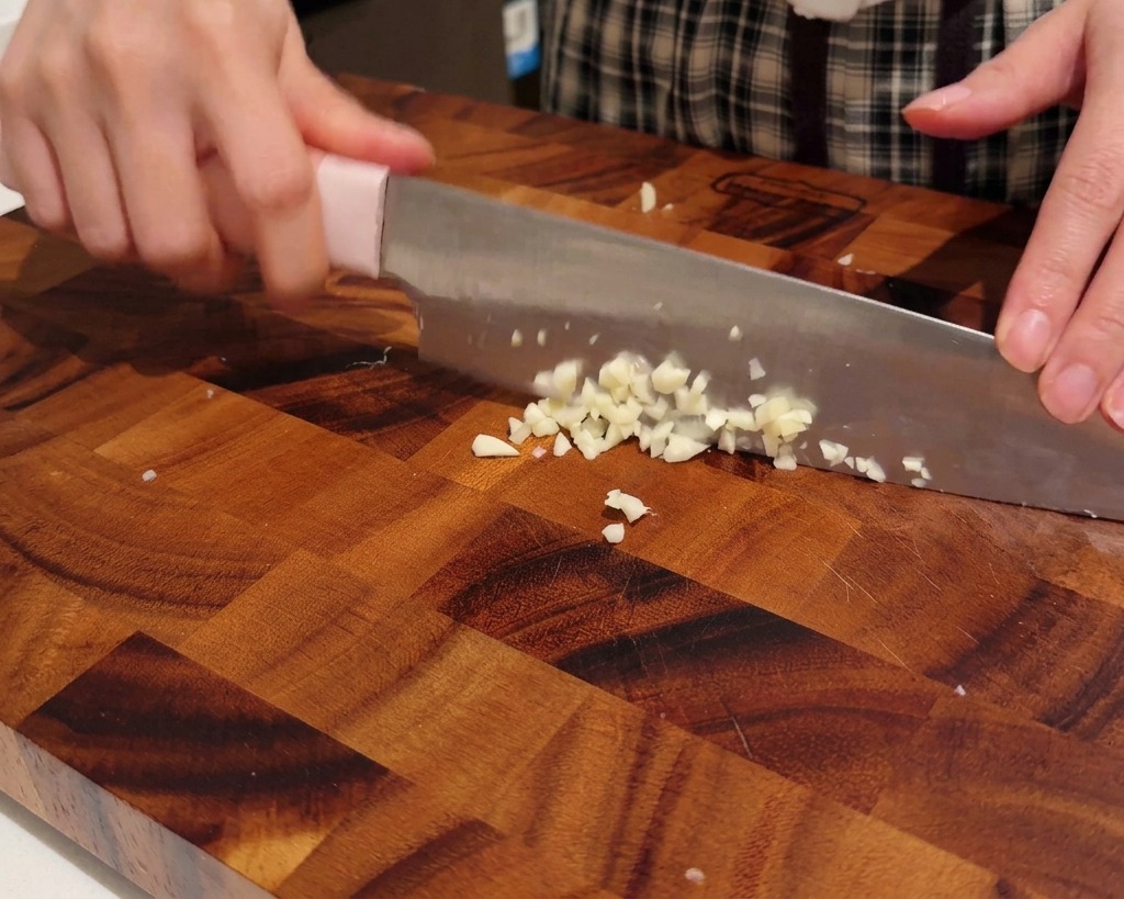 Mani che tritano spicchi daglio finemente su un tagliere di legno.
