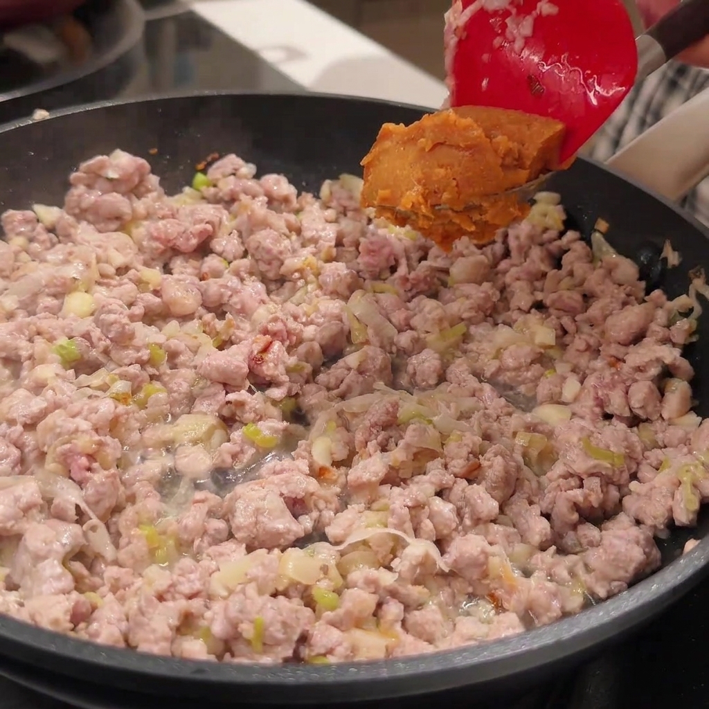 Una spatola rossa che aggiunge pasta di miso sopra il maiale rosolato in padella.