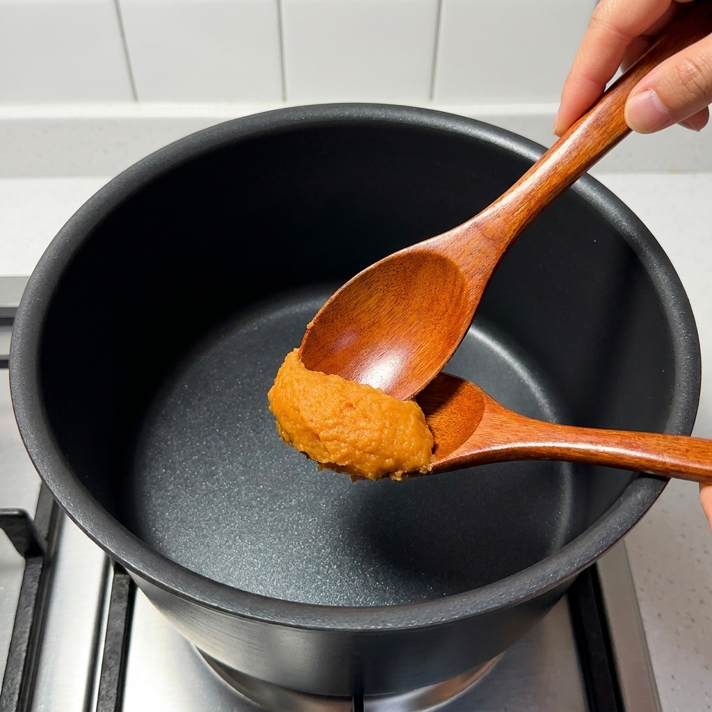 Un cucchiaio di legno mescola miso, mirin, zucchero, brodo di ossa di maiale e succo di yuzu in un pentolino sul fornello.