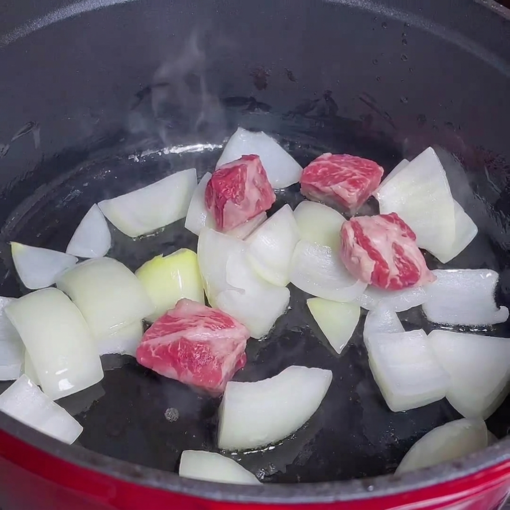 Cubetti di carne che cuociono insieme alle cipolle in una pentola rossa.