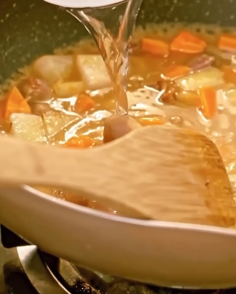 Acqua versata in una padella piena di manzo, verdure e curry che si scioglie, con una spatola di legno visibile.