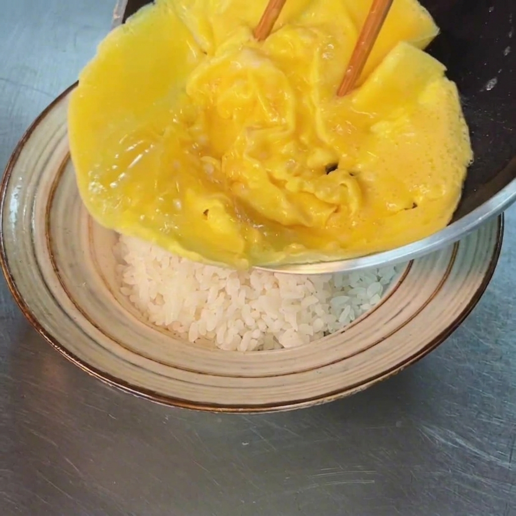 Lomelette soffice che viene fatta scivolare dalla padella su una ciotola di riso bianco.