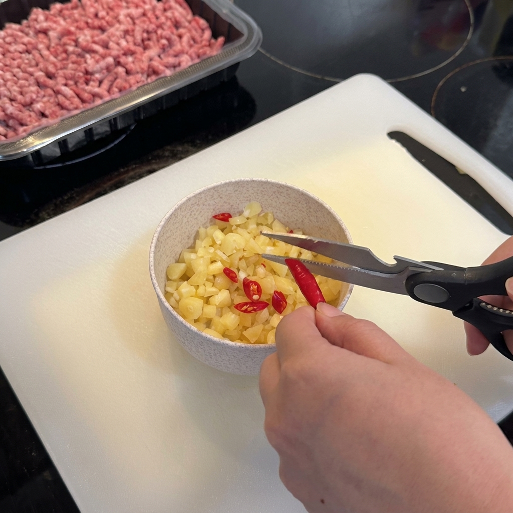 Forbici da cucina che tagliano peperoncini rossi vivaci birds eye in una ciotola contenente aglio tritato.