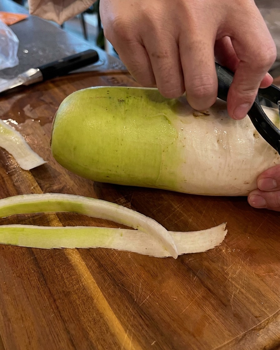 Mani che usano un pelapatate nero per rimuovere la buccia verde e bianca da un grande ravanello su un tagliere di legno.