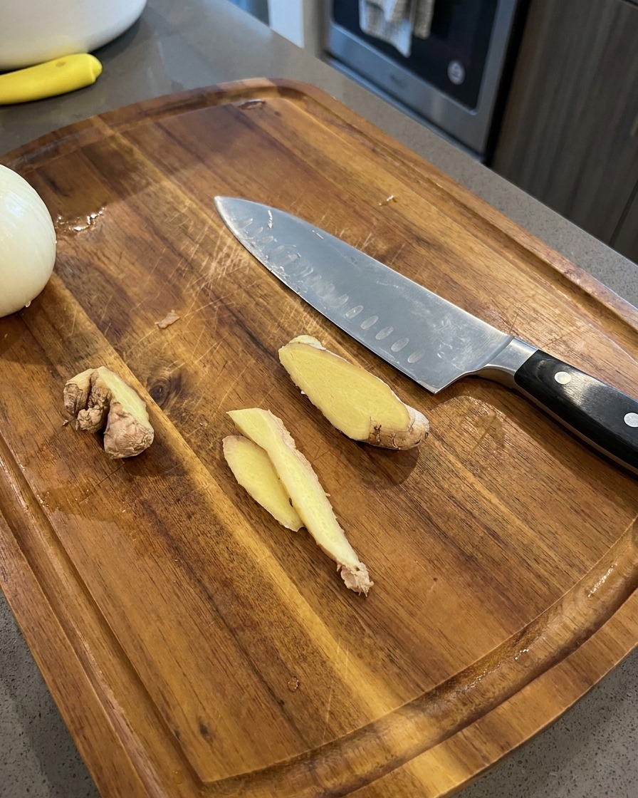 Fette sottili di zenzero fresco su un tagliere di legno accanto a un coltello da chef e una metà di cipolla.
