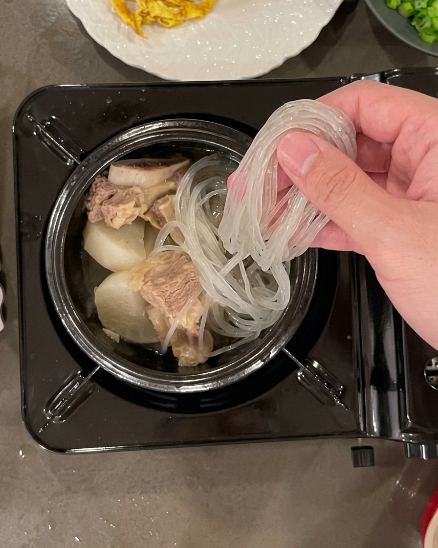 Una mano che mette un fascio di noodles di fecola ammollati e traslucidi in una pentola di pietra riempita con carne e pezzi di ravanello.