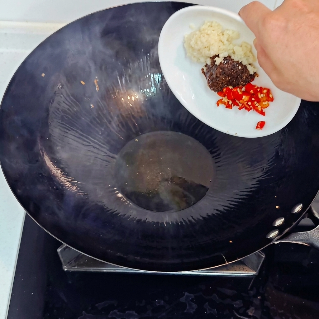 Aggiunta di aglio tritato, peperoncino rosso tritato e pasta di soia in un wok caldo da un piattino bianco.