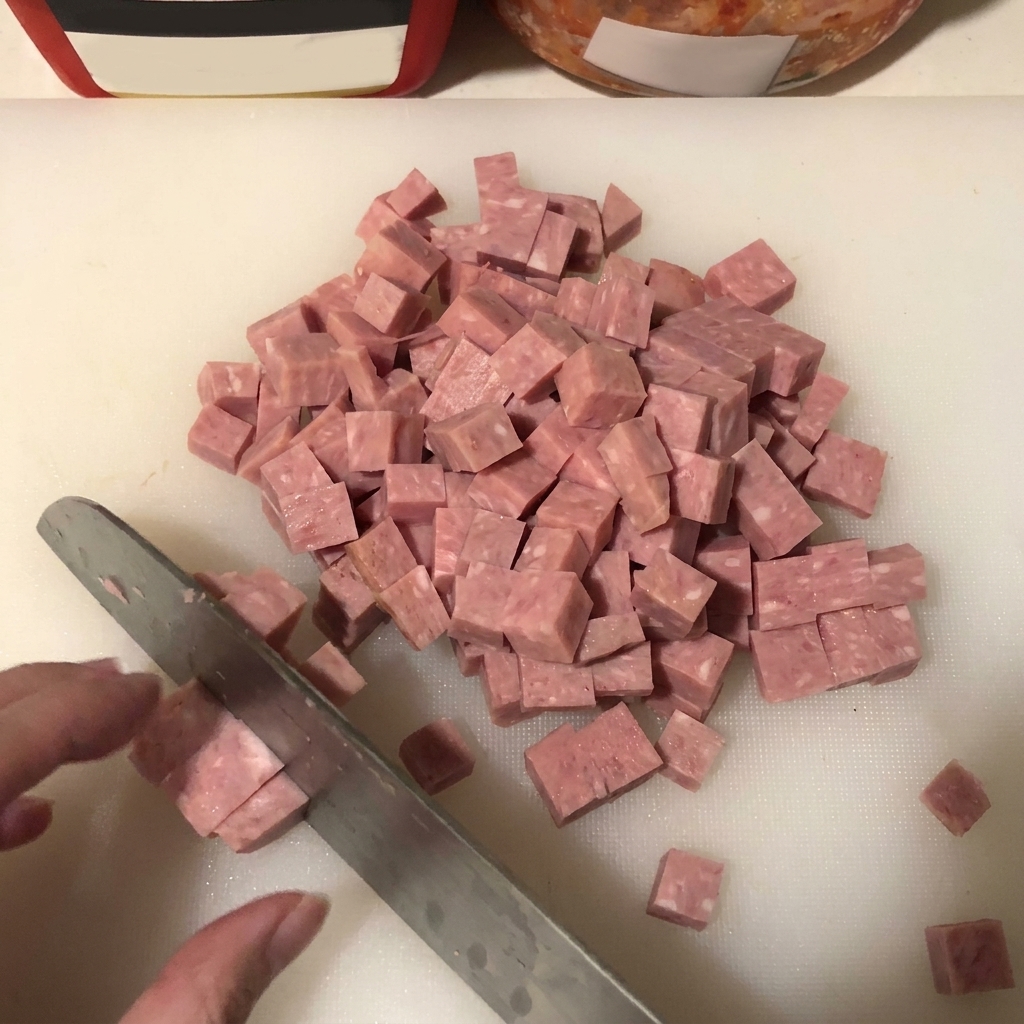 Primo piano di spam tagliato a cubetti uniformi su un tagliere bianco con una lama di coltello visibile.