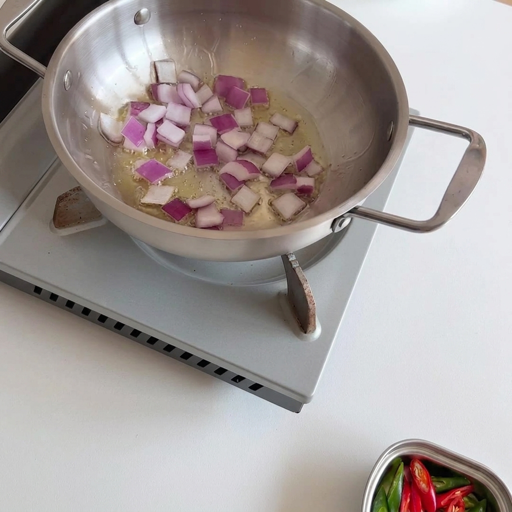 Cipolle rosse a dadini che sfrigolano e si ammorbidiscono in olio caldo in una padella di acciaio inossidabile, mescolate con un cucchiaio di legno.