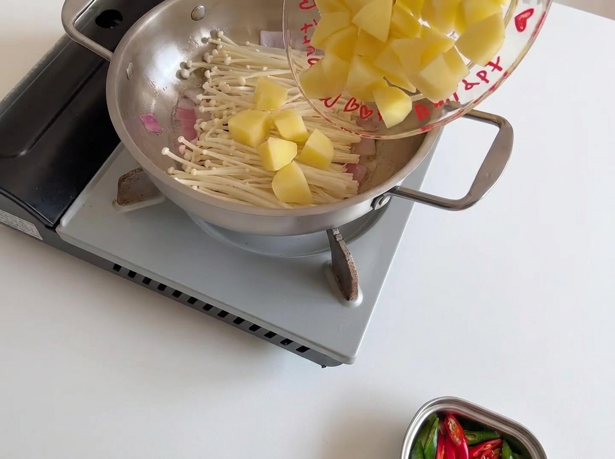 Pezzi di patata gialla cruda aggiunti nella padella, appoggiati sopra i funghi enoki bianchi.