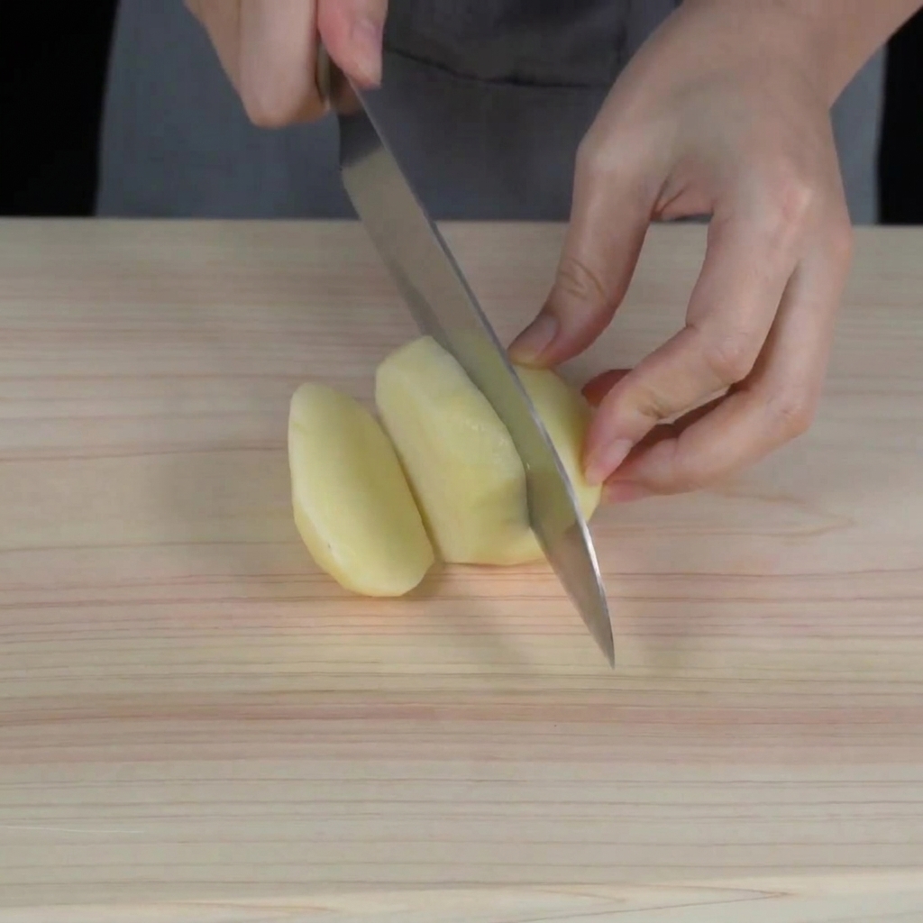 Mani che usano un coltello da chef per tagliare una patata cruda sbucciata a cubetti su un tagliere di legno.