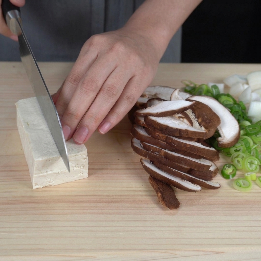Mani che tagliano un panetto di tofu bianco solido in cubetti uniformi su un tagliere.