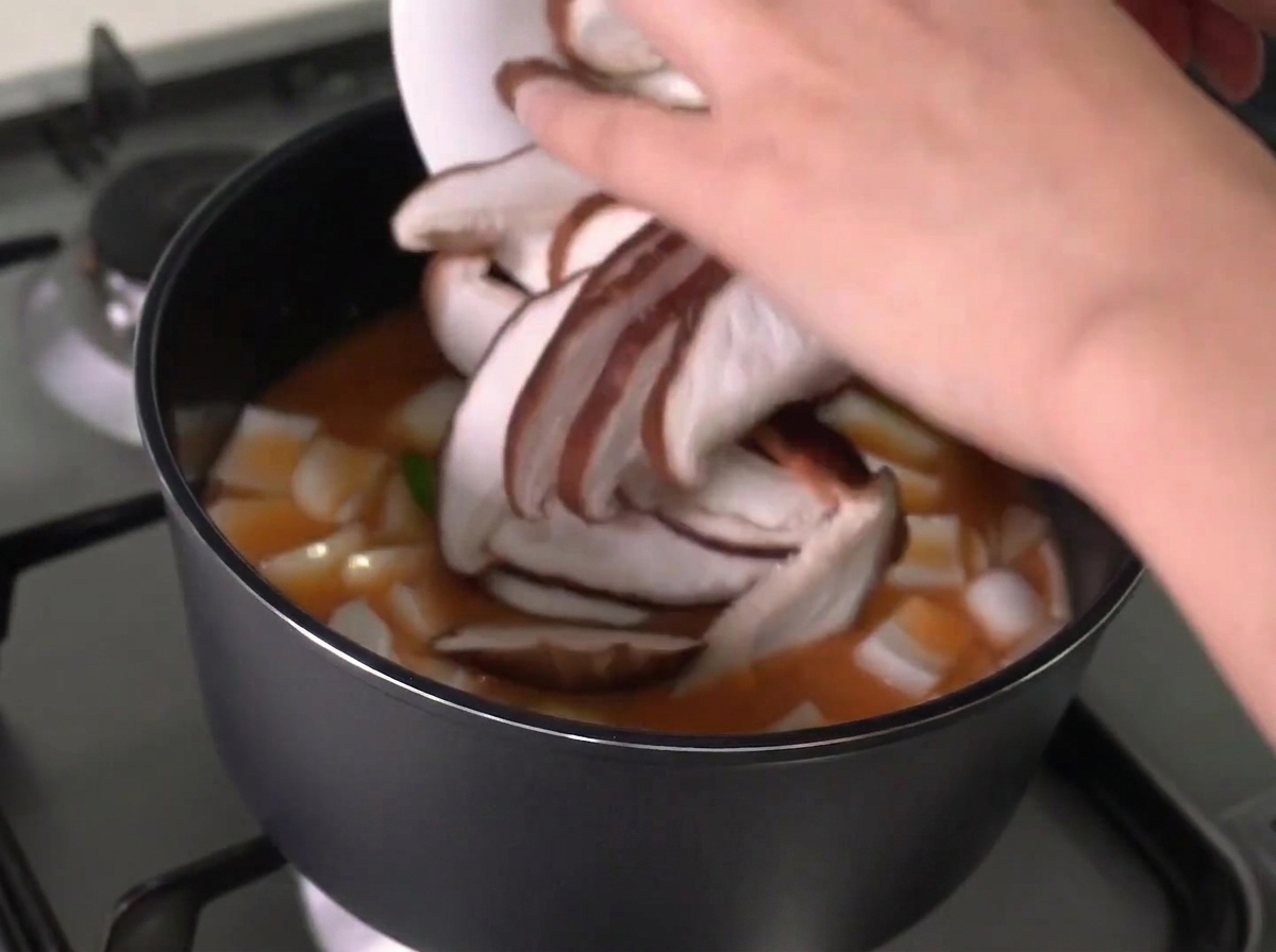 Funghi shiitake affettati aggiunti da un piatto bianco in una pentola di stufato bollente.