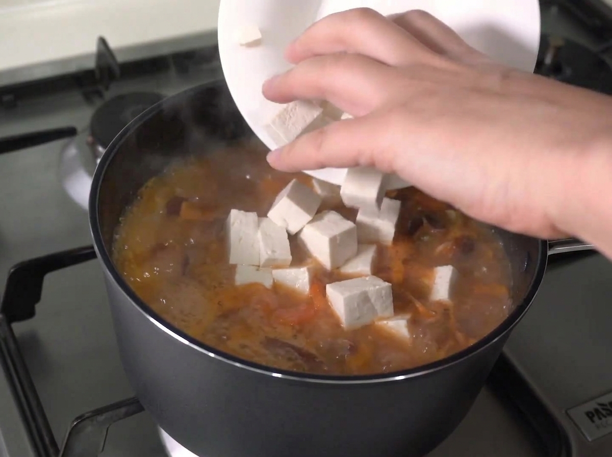 Cubetti di tofu fresco aggiunti da un piatto bianco in uno stufato in ebollizione attiva.