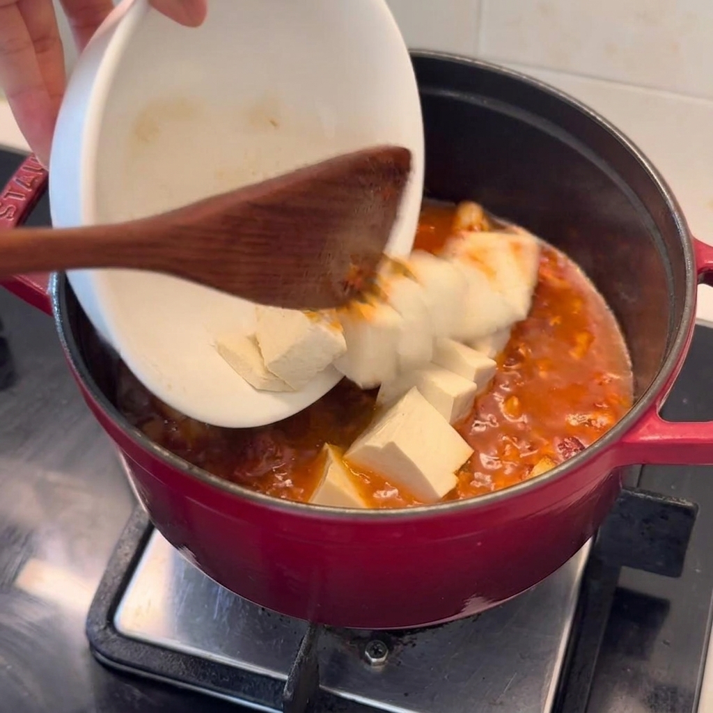 Cubetti di tofu fresco aggiunti con cura da una ciotola bianca in una pentola rossa ribollente.