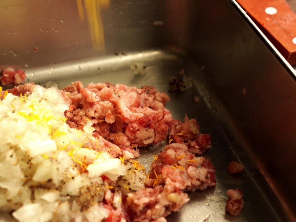Cipolla tritata e pangrattato aggiunti alla carne macinata cruda in un contenitore metallico.