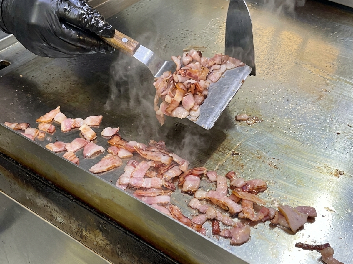 Una spatola di metallo che mescola pezzi di bacon su una piastra calda per sciogliere il grasso.