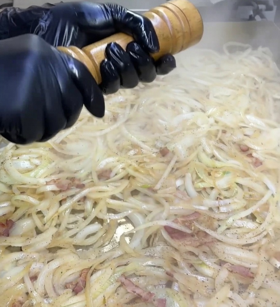 Mani con guanti neri che girano un macinapepe in legno sopra una piastra coperta di cipolle e bacon in cottura.
