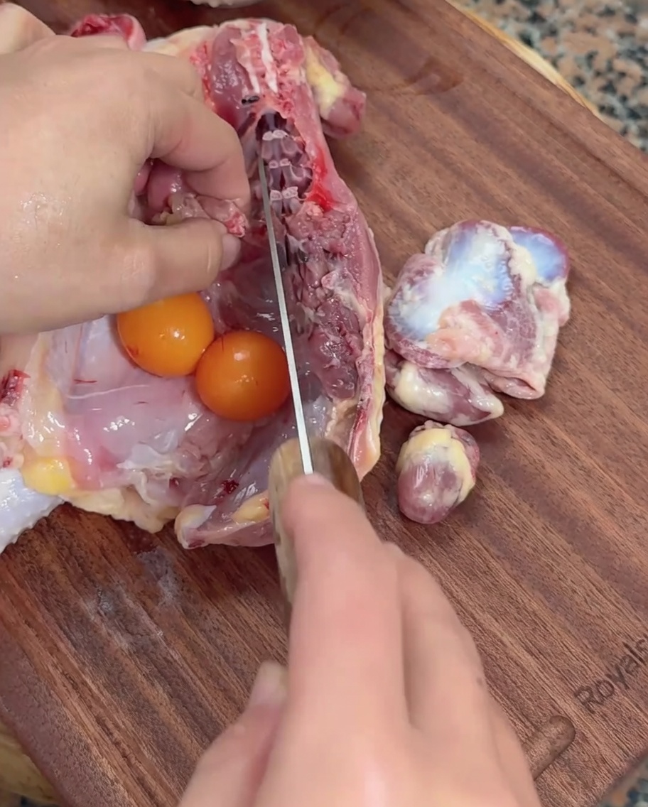 Mani che usano una mannaia per incidere le frattaglie di pollo crudo accanto a uova di gallina su un tagliere di legno.