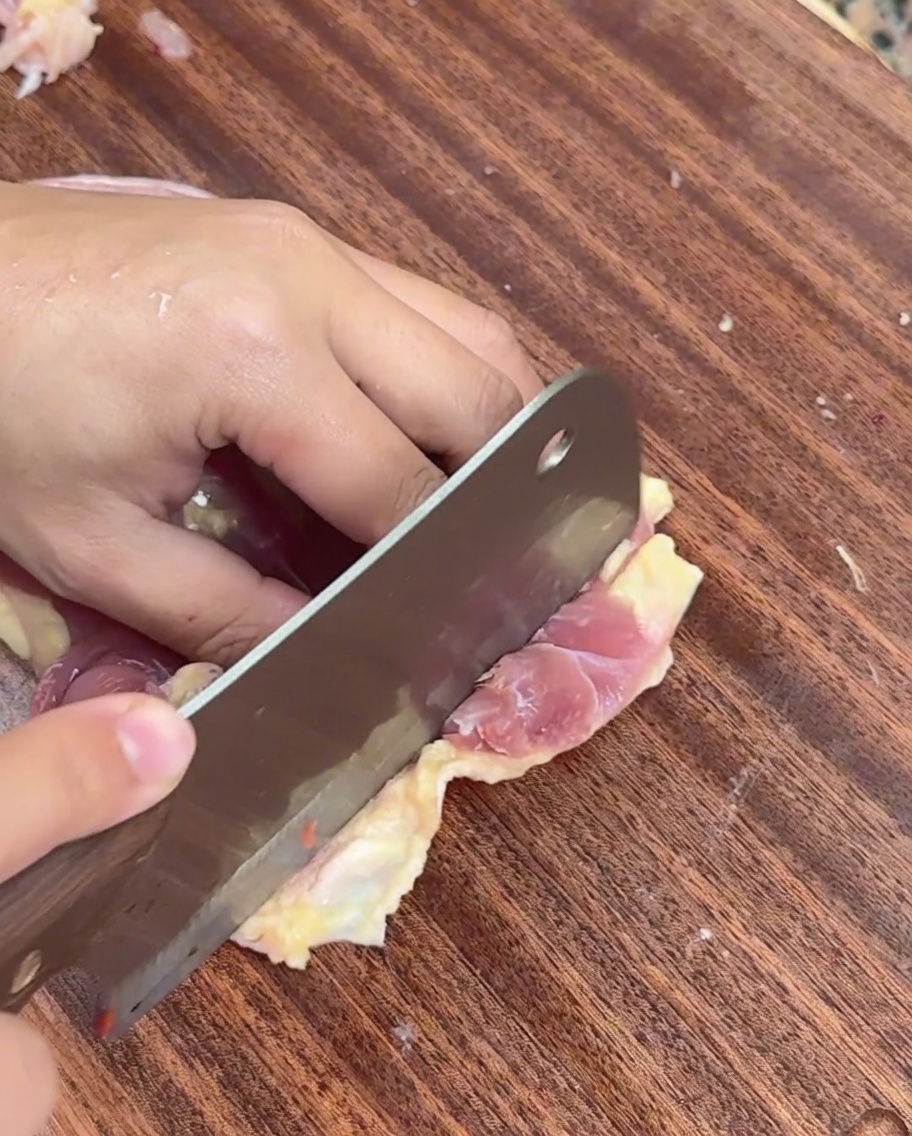 Una mano che usa una mannaia per affettare la carne di pollo cruda in strisce sottili su un tagliere.