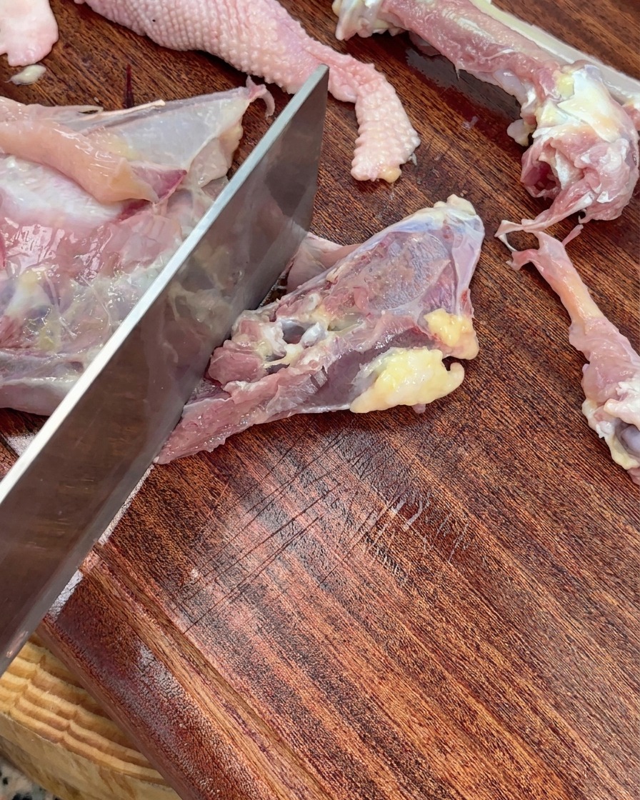 Una mannaia che trita lo scheletro di un pollo crudo in grossi pezzi su un tagliere di legno.