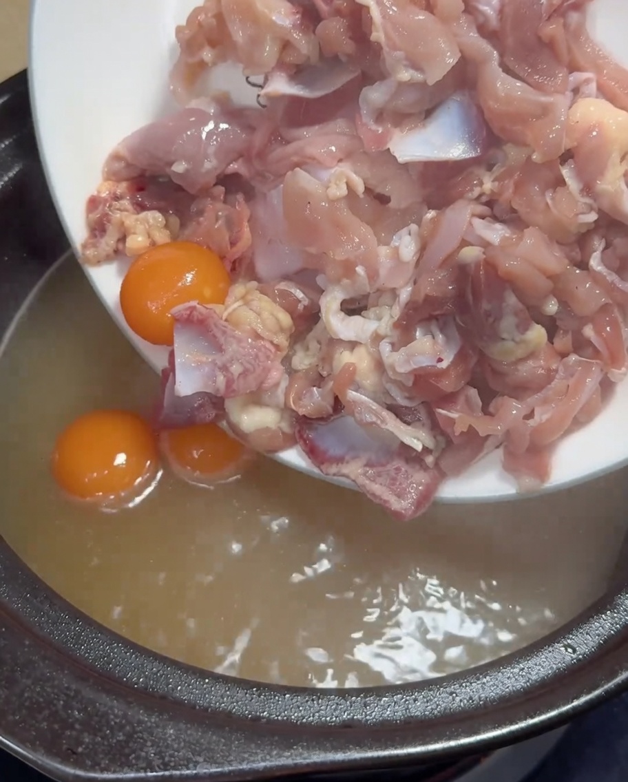 Carne di pollo cruda affettata sottilmente e tuorli gialli aggiunti in una pentola di brodo di pollo dorato bollente.