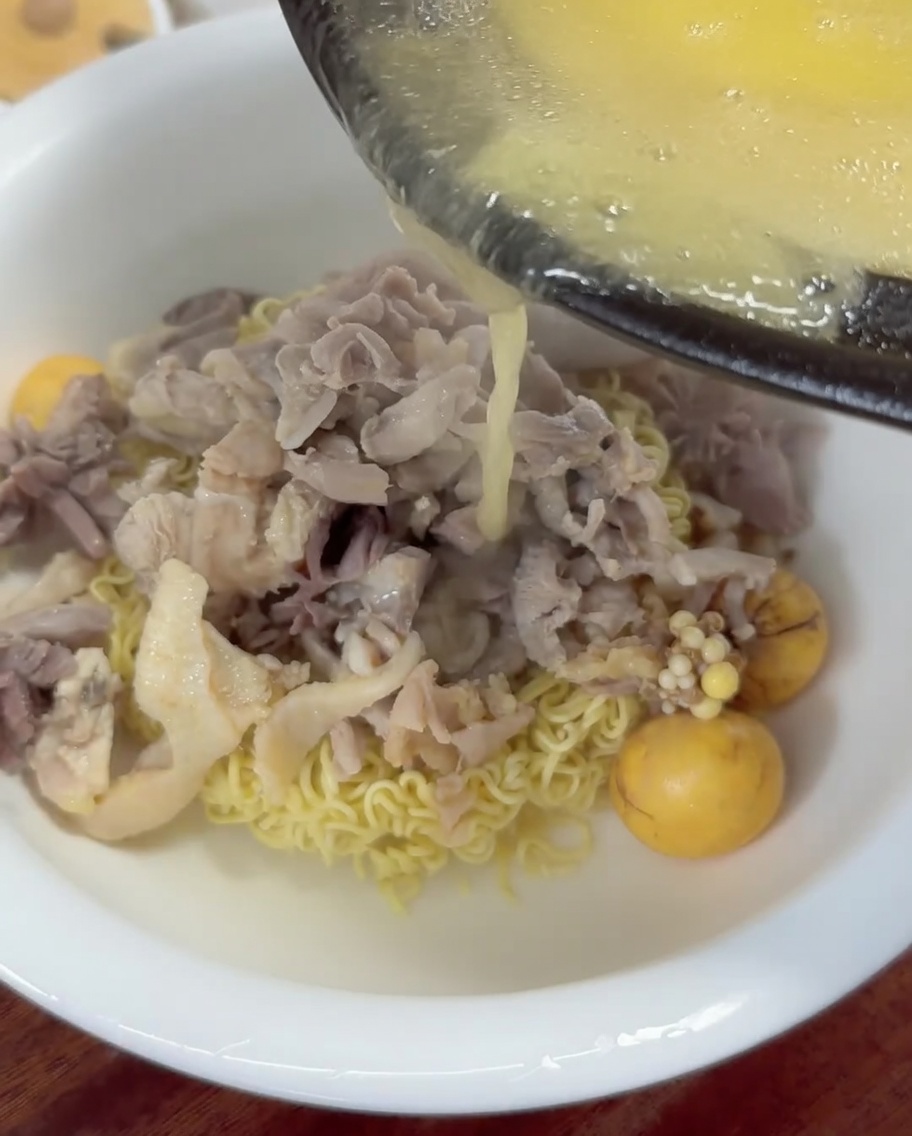 Brodo di pollo dorato caldo che viene versato da un mestolo in una ciotola di noodles e pollo.