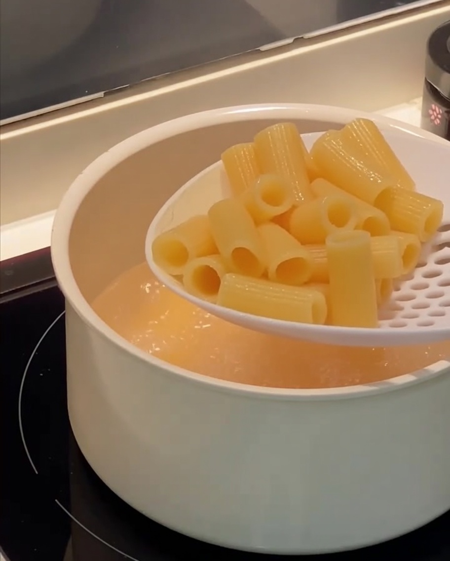 Una schiumarola che solleva i rigatoni cotti da una pentola di acqua bollente.